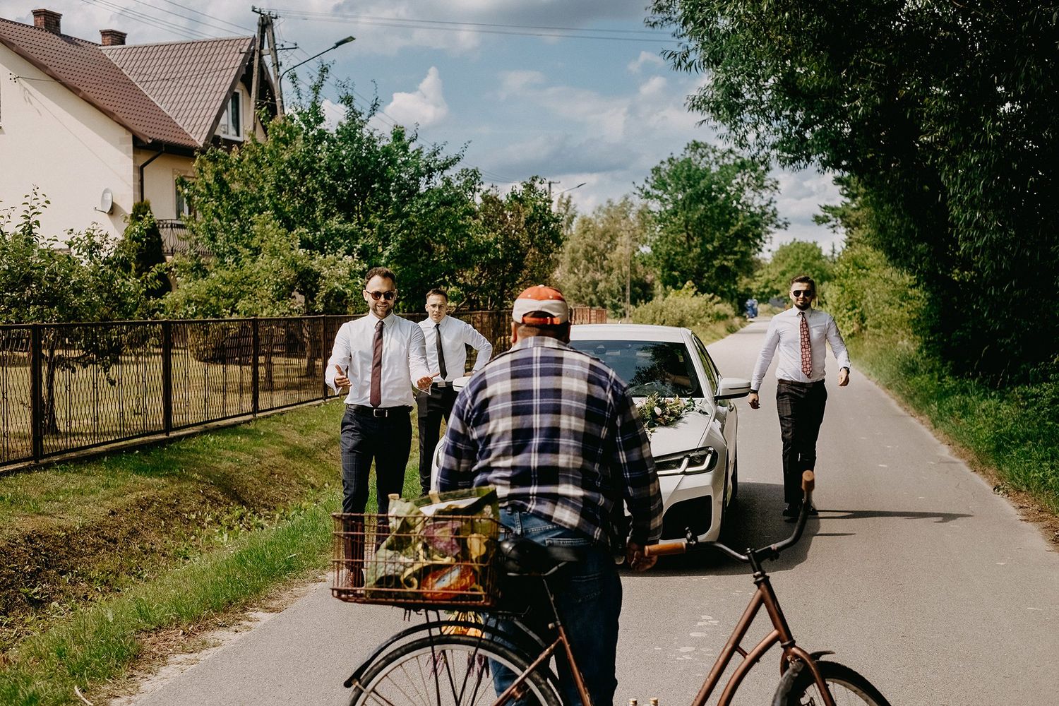Wedding party in formal attire walking down a rural road past a cyclist on a sunny day.