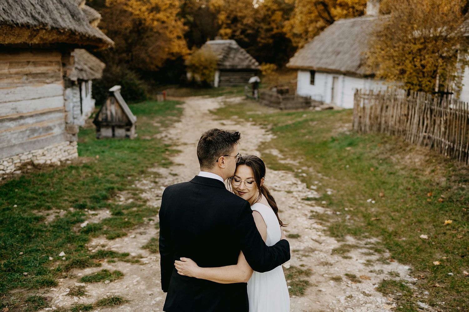 Couple embracing in a rustic autumn village setting with traditional wooden cottages and fall foliage.