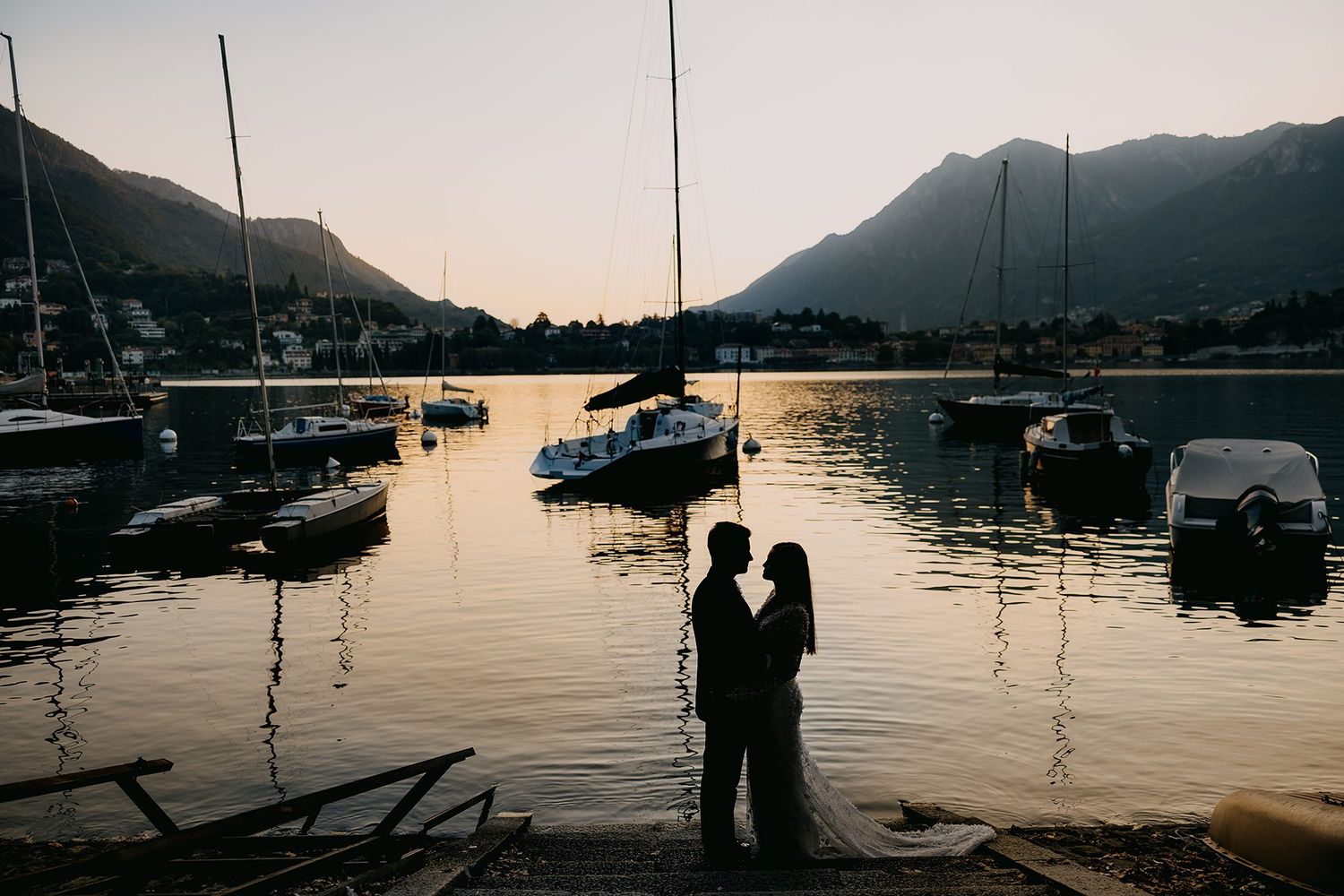 Silhouette of wedding couple standing romantically at sunset harbor with sailboats and mountain backdrop.