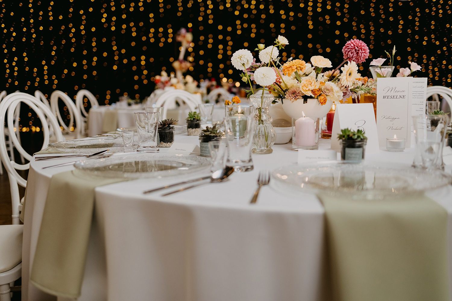 Elegant wedding reception table set with floral centerpieces, candles, and string lights in the background.