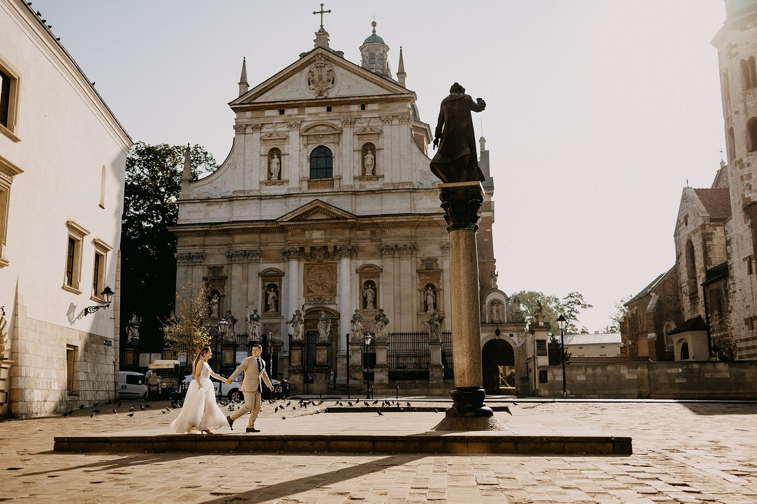 Bride and groom walk hand in hand in a historic European square with a baroque church and statue backdrop.