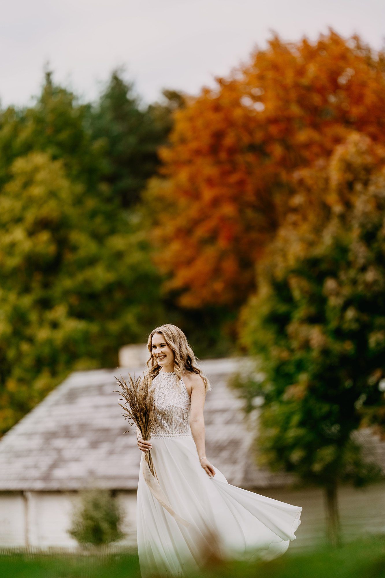 Bride in white gown holding dried bouquet walks through autumn garden with colorful fall foliage.