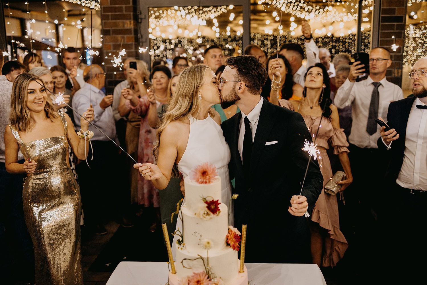 Bride and groom kiss beside wedding cake while guests celebrate with sparklers at reception.