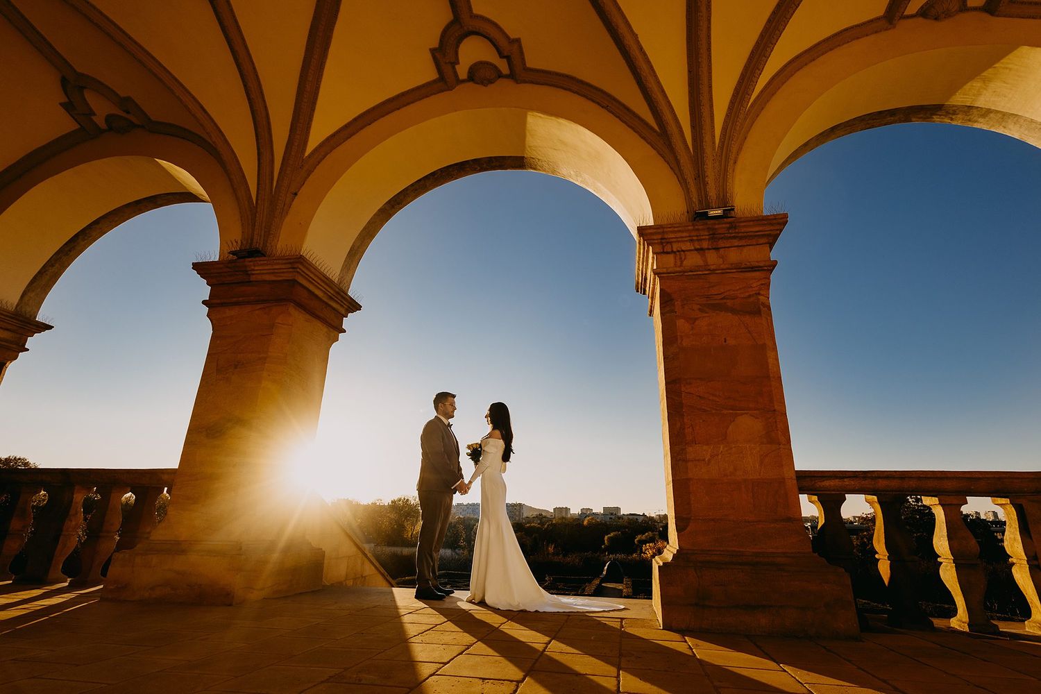 Bride and groom holding hands under stone arches at golden sunset.