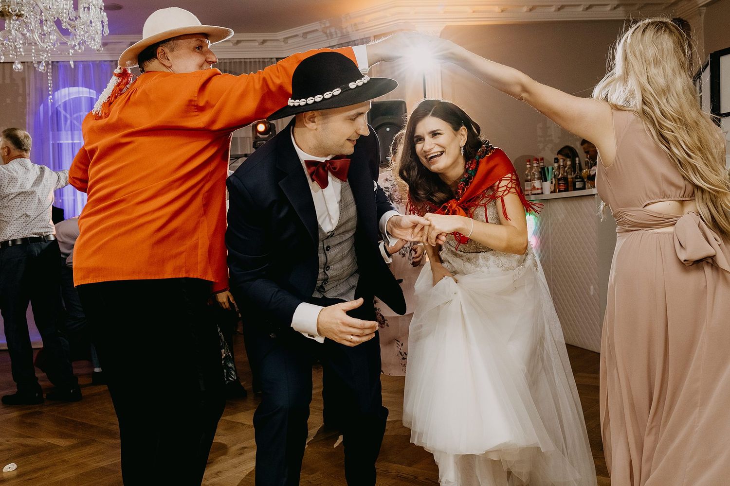 Bride and groom dancing with guests wearing colorful outfits and hats at a lively wedding reception.