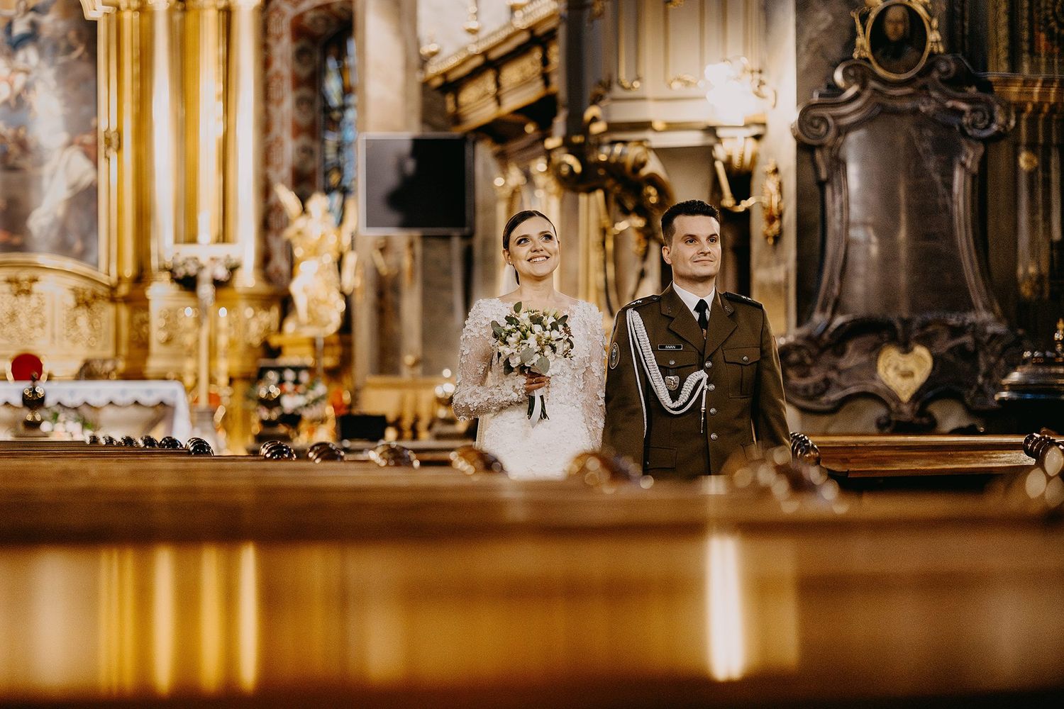 A bride and groom stand together at the ornate golden altar of a baroque church during their wedding ceremony.