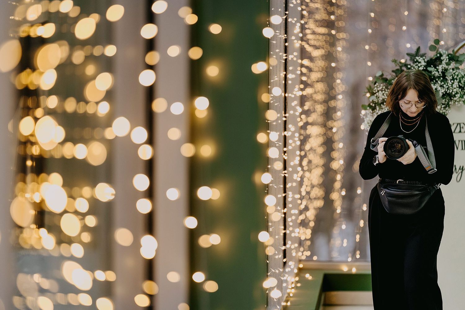 Woman in black coat holding camera amid stunning bokeh fairy light curtains at event.