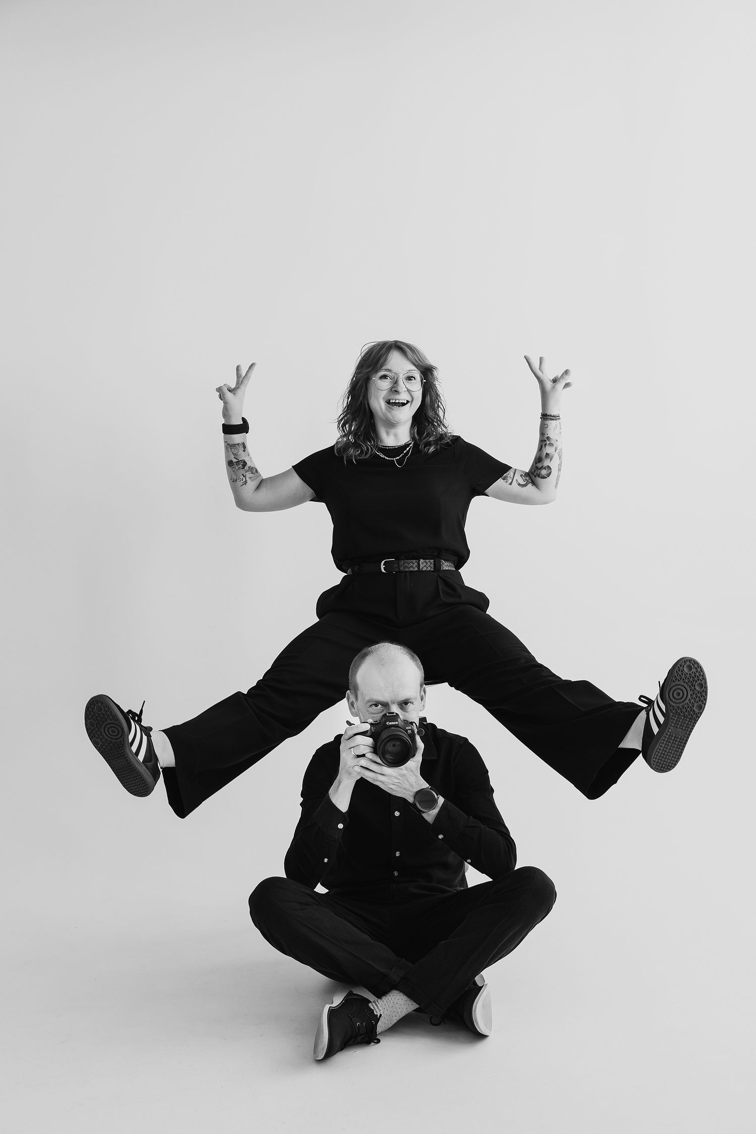 Black and white photo of two people in black outfits, woman posing above seated man holding camera.