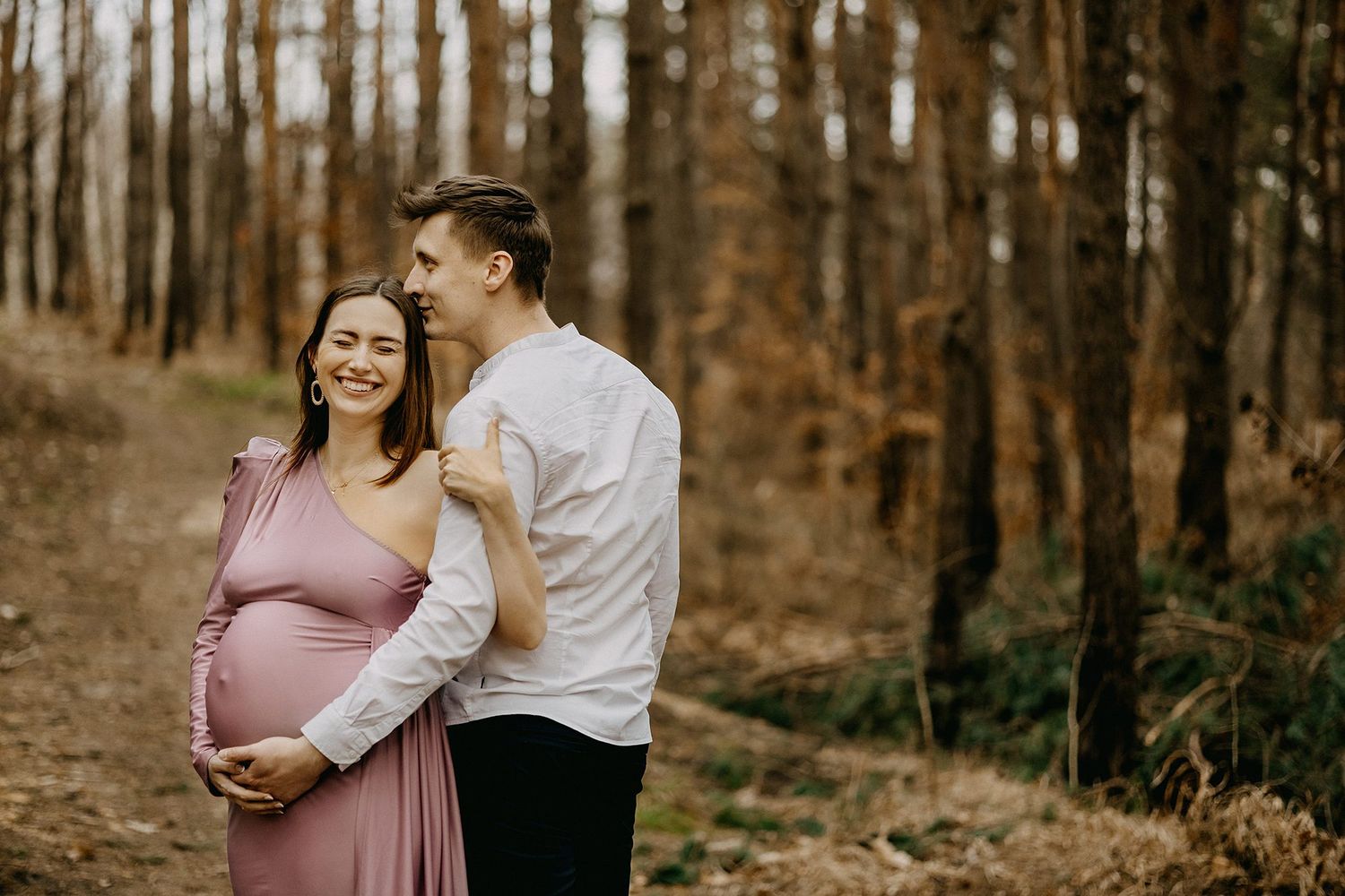 Couple embracing in autumn pine forest, pregnant woman in pink dress smiling joyfully.
