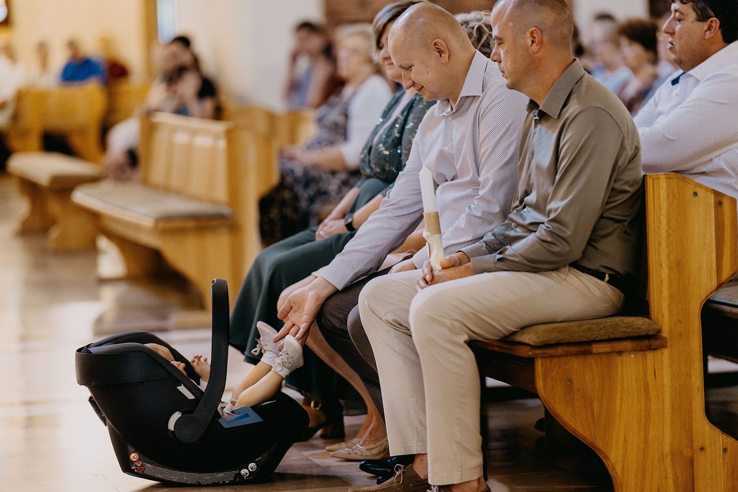 Two men sitting in church pew with infant in car seat during baptism service.