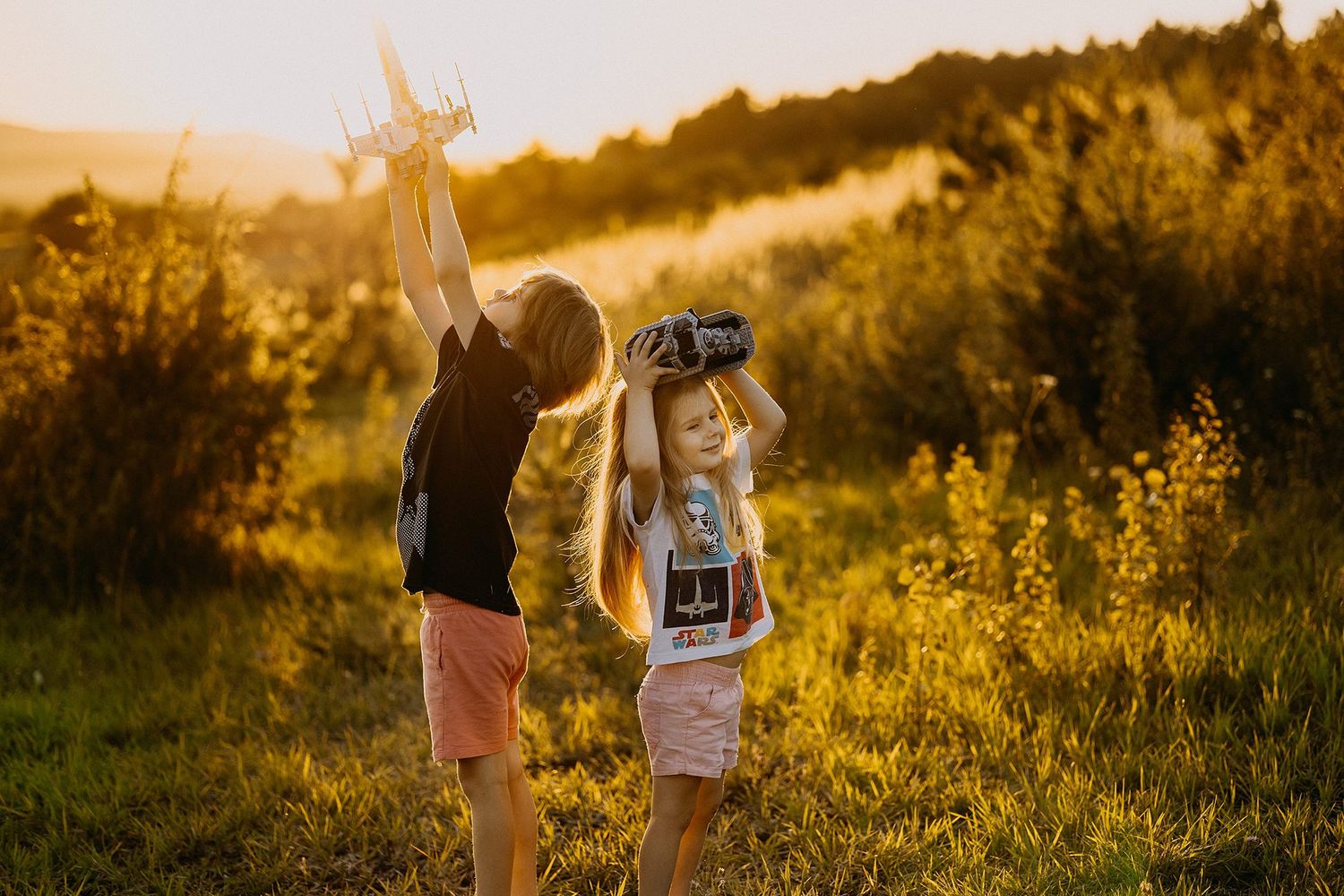 Two children playing outdoors at sunset, one holding toy airplane and other using VR headset.