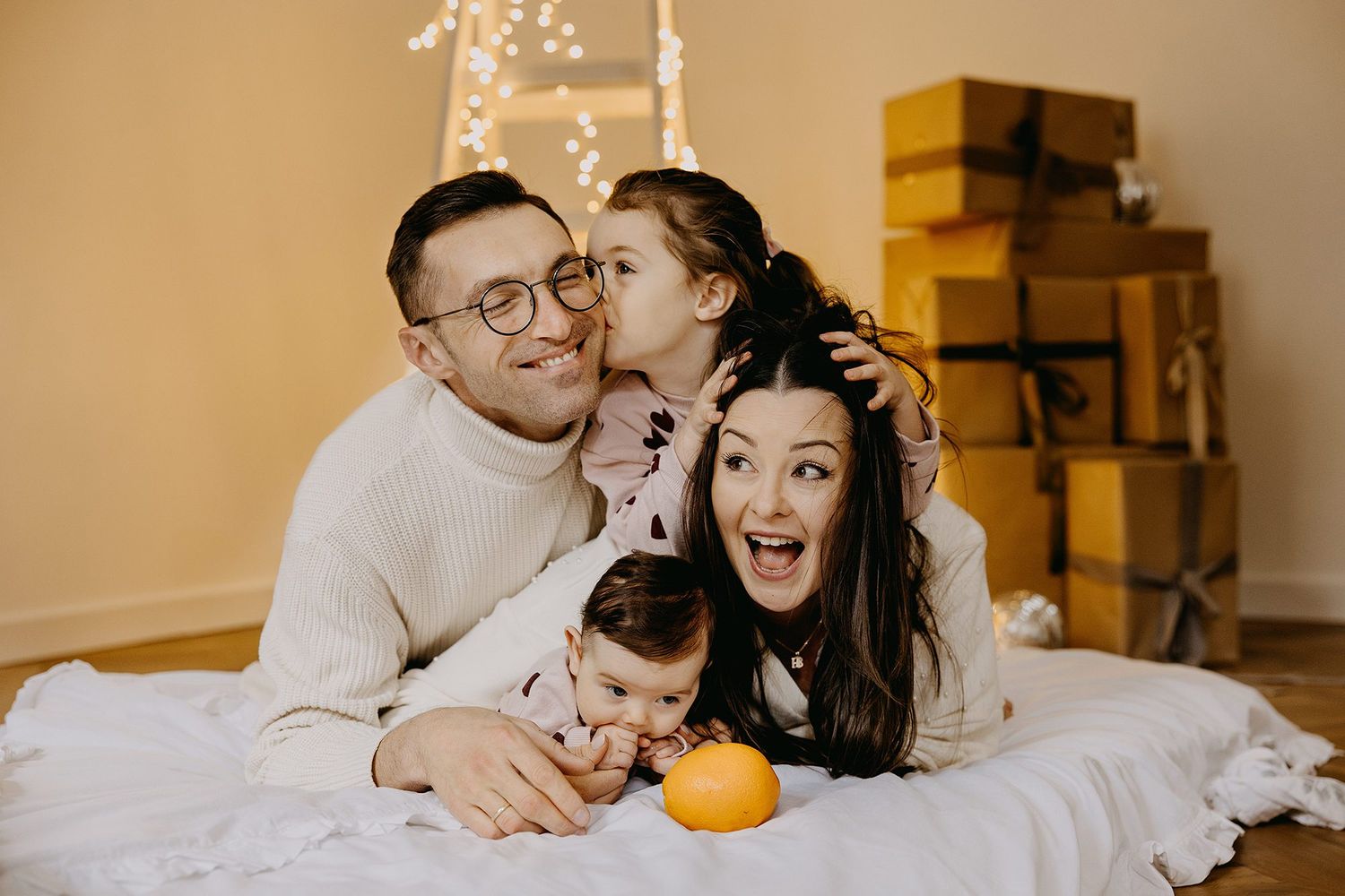 A happy family of four laughs and cuddles together on the floor surrounded by golden Christmas gift boxes.