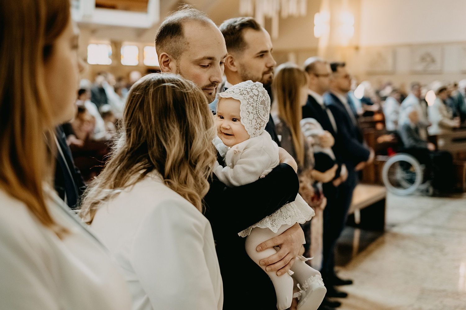 Father proudly holds smiling baby in white bonnet during Catholic baptism church ceremony.