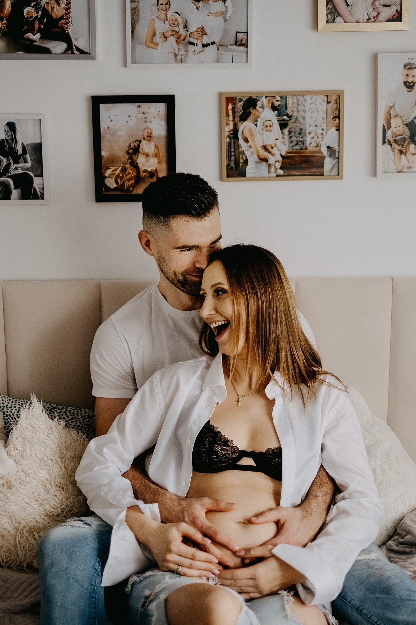 Happy pregnant couple sitting on bed, man embracing woman showing baby bump, photo gallery wall behind them.