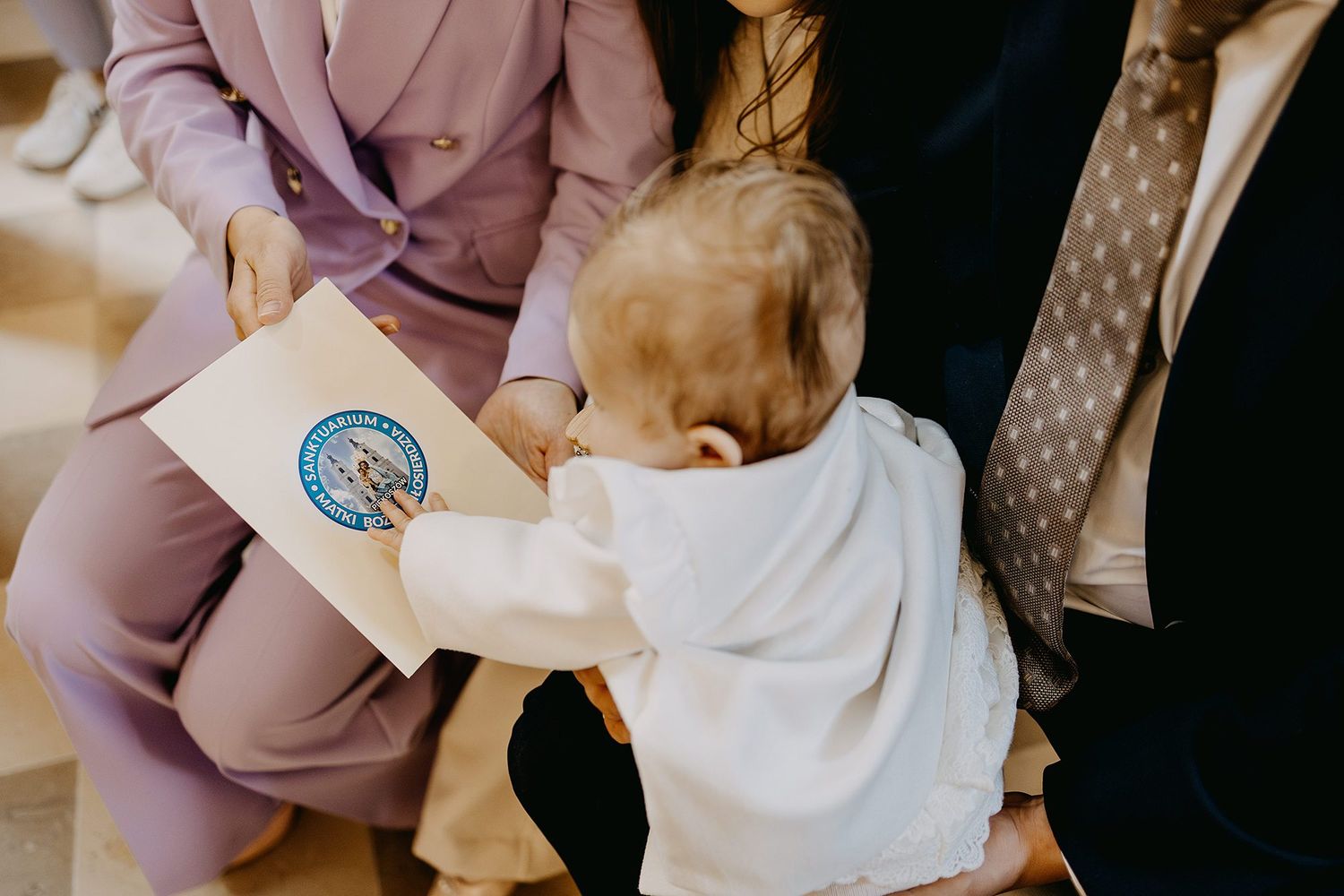 A baby in white christening outfit reaches for a baptism certificate held by parents.