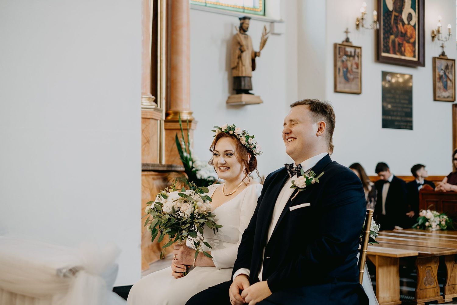 Bride with floral crown and groom in tuxedo smiling during church wedding ceremony.