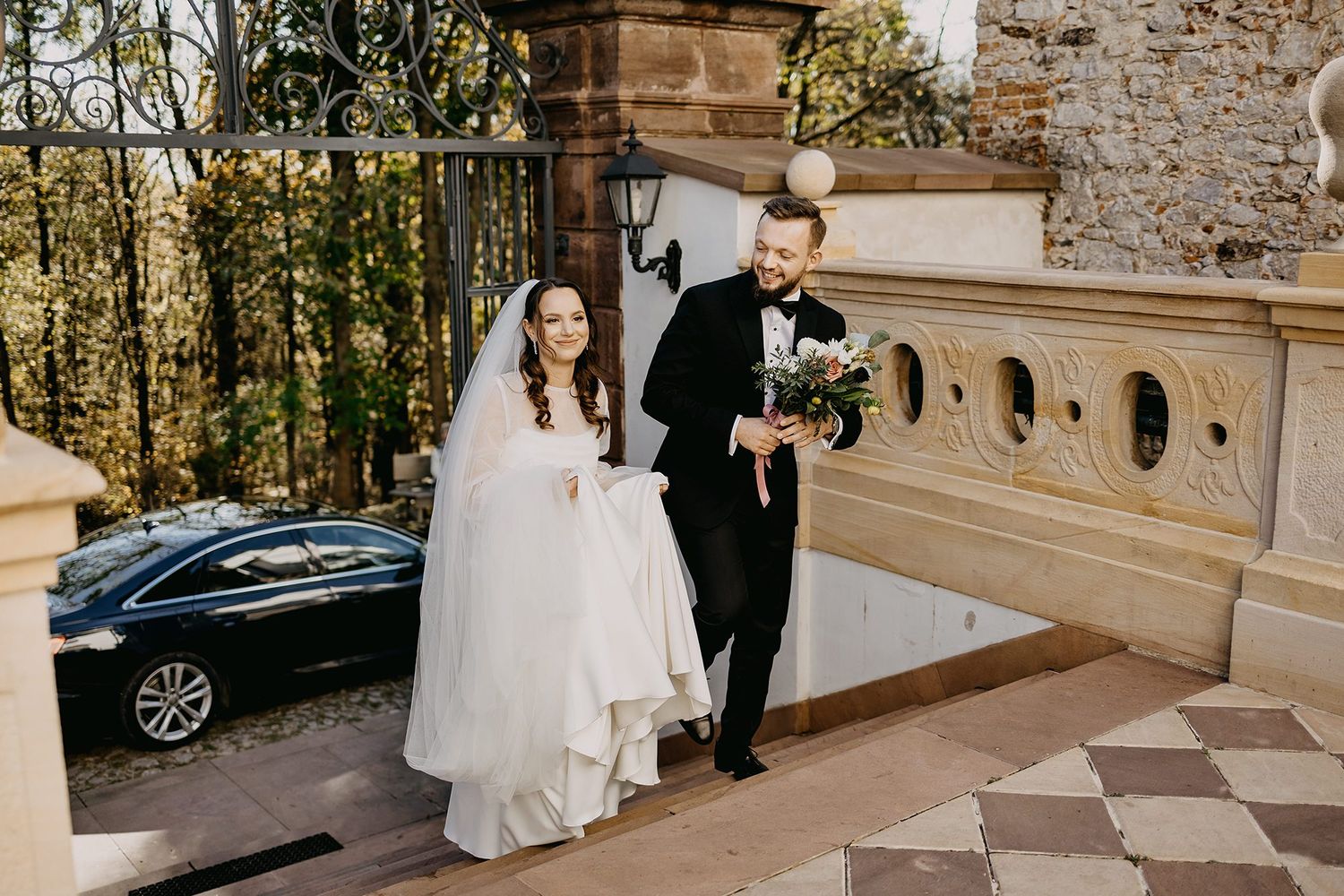 Bride and groom in formal attire walk down stone steps outside a historic venue with autumn trees.