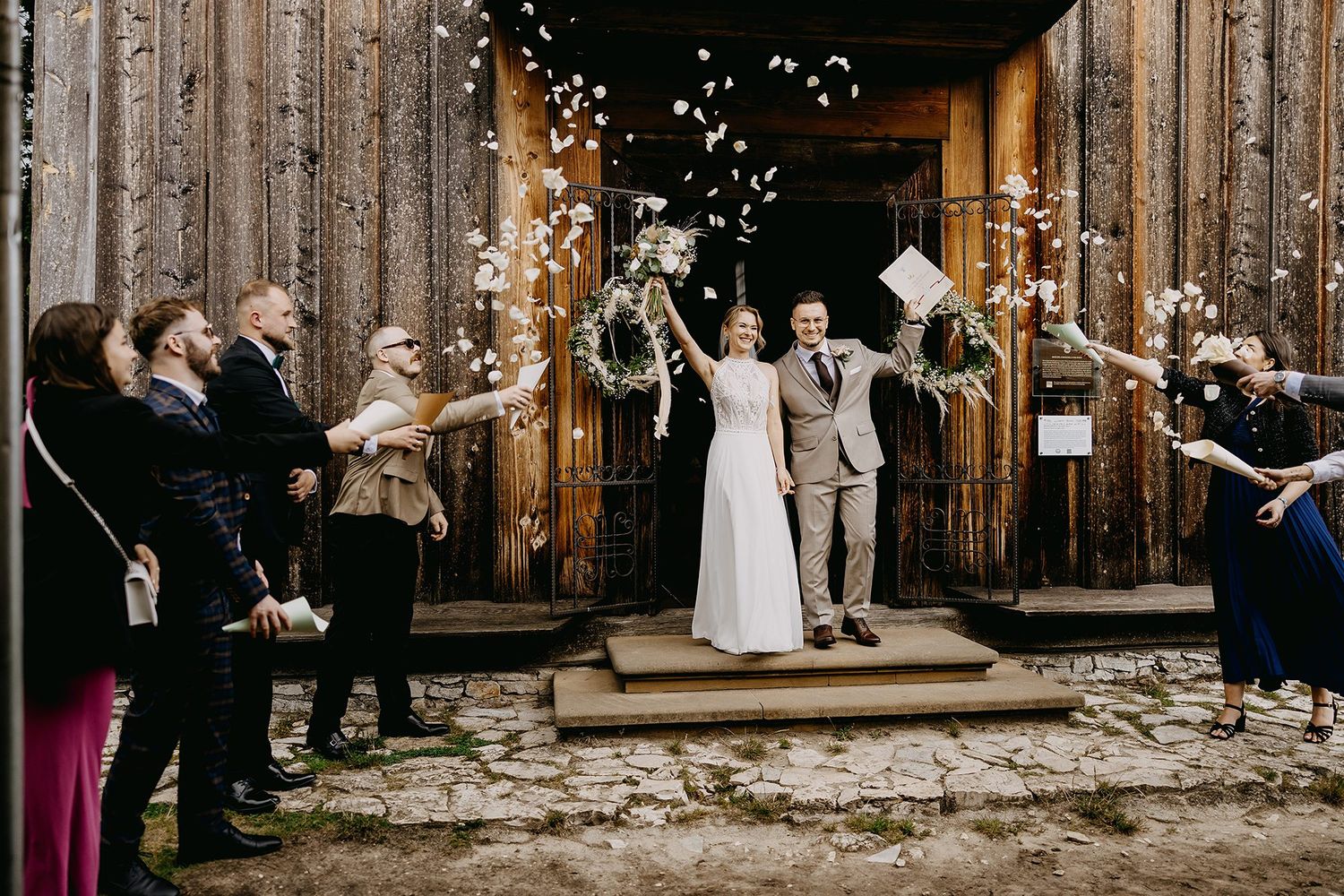 Bride and groom exit rustic wooden barn doors as guests shower them with white flower confetti.