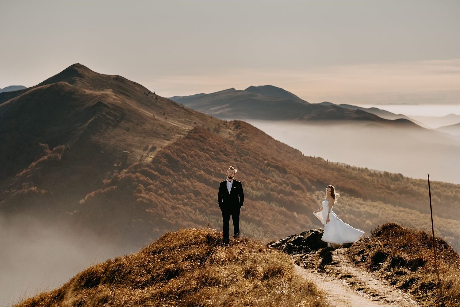Bride and groom standing on a mountain ridge with dramatic fog-covered valleys and peaks behind them.