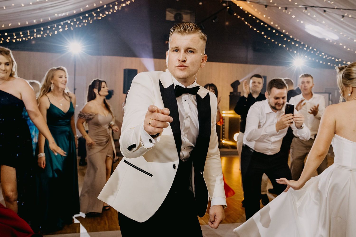 Groom in white tuxedo jacket dancing energetically at a lively wedding reception with guests.
