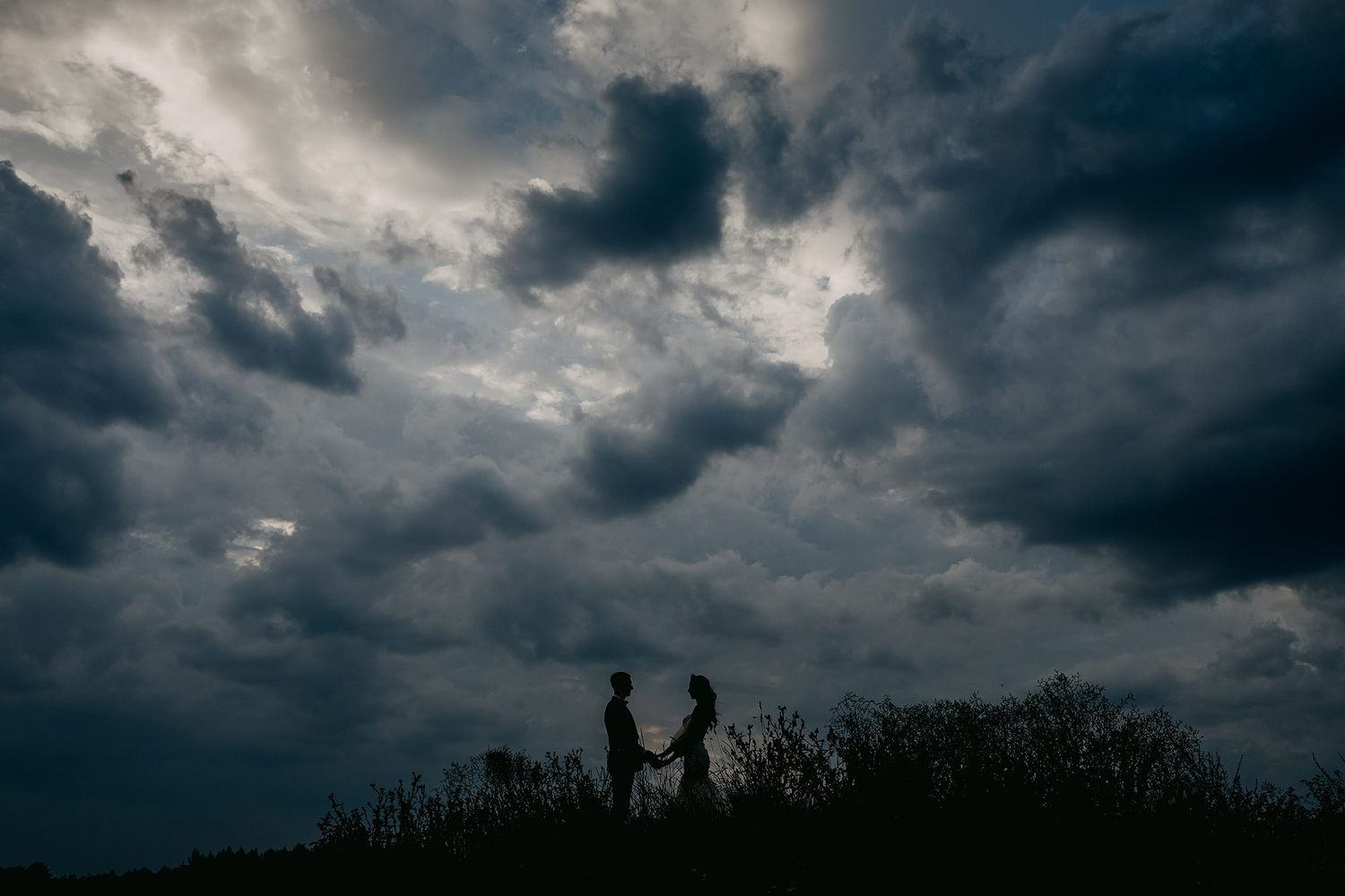 Silhouette of a couple holding hands on a hilltop beneath dramatic stormy clouds.