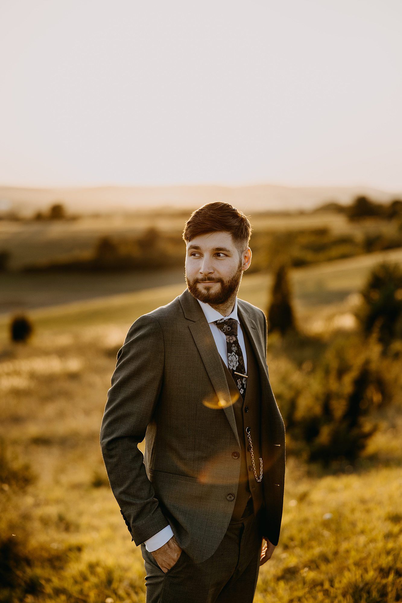 Handsome bearded groom in dark suit posing confidently in a golden sunset countryside landscape.