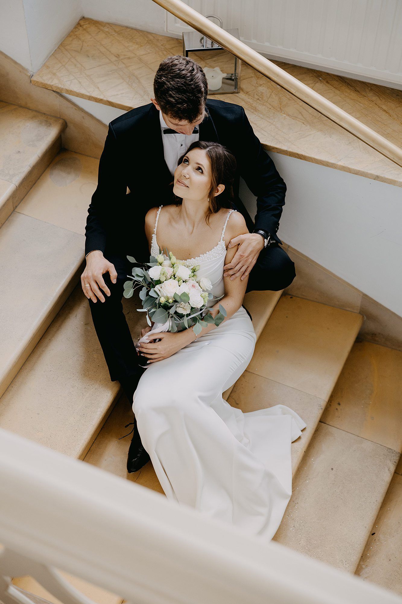 Bride and groom sitting on marble stairs, bride holding white bouquet while groom embraces her.