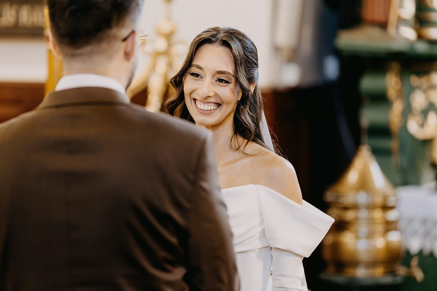 Radiant bride in off-shoulder white gown smiles beautifully at groom during church wedding ceremony.