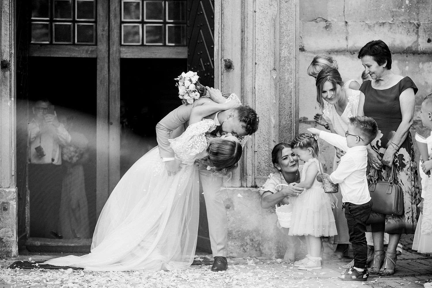 Bride bends down to embrace children outside church doors in charming black and white wedding photo.