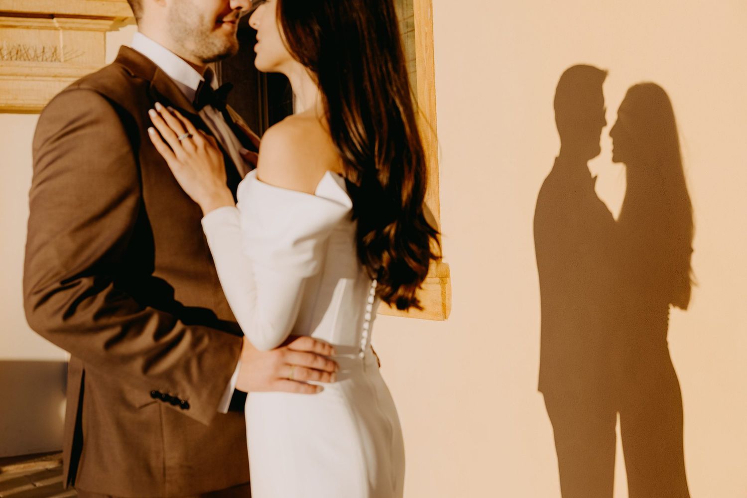 Couple shares romantic moment in wedding attire with silhouette shadow on warm wall behind them.