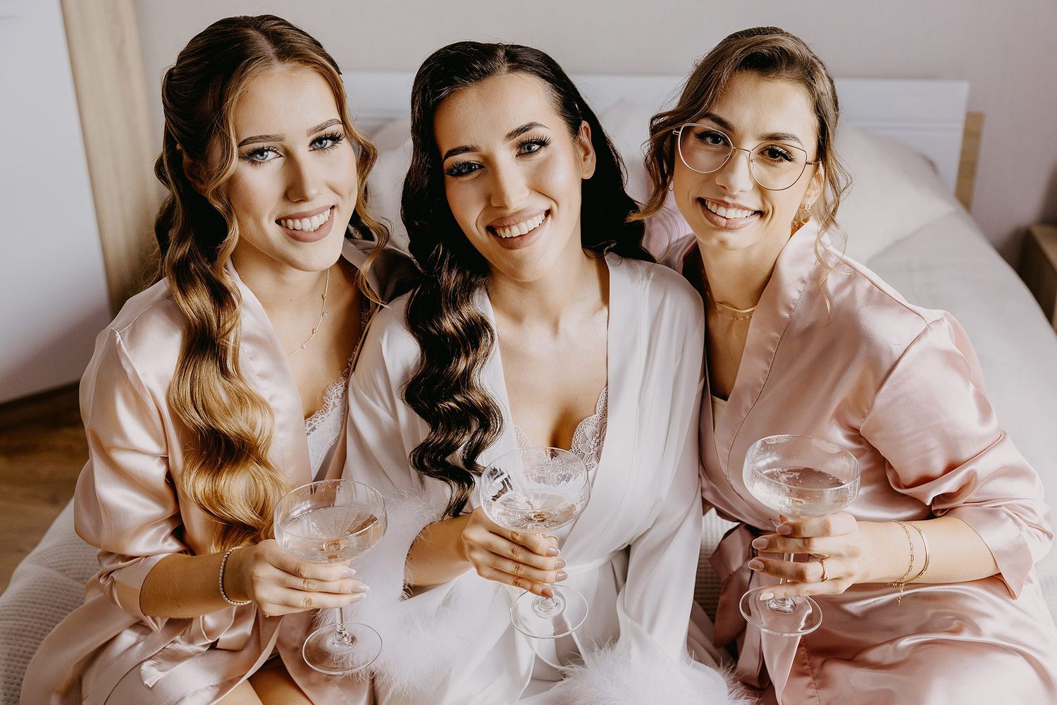 Three bridesmaids in pink satin robes smiling and holding champagne glasses.