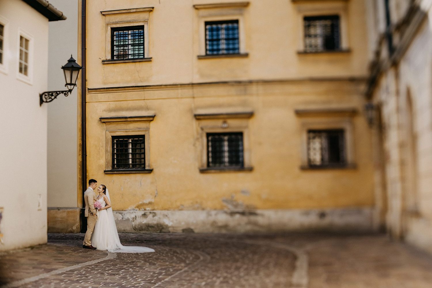 Wedding couple embraces beside a historic yellow building on a cobblestone street.