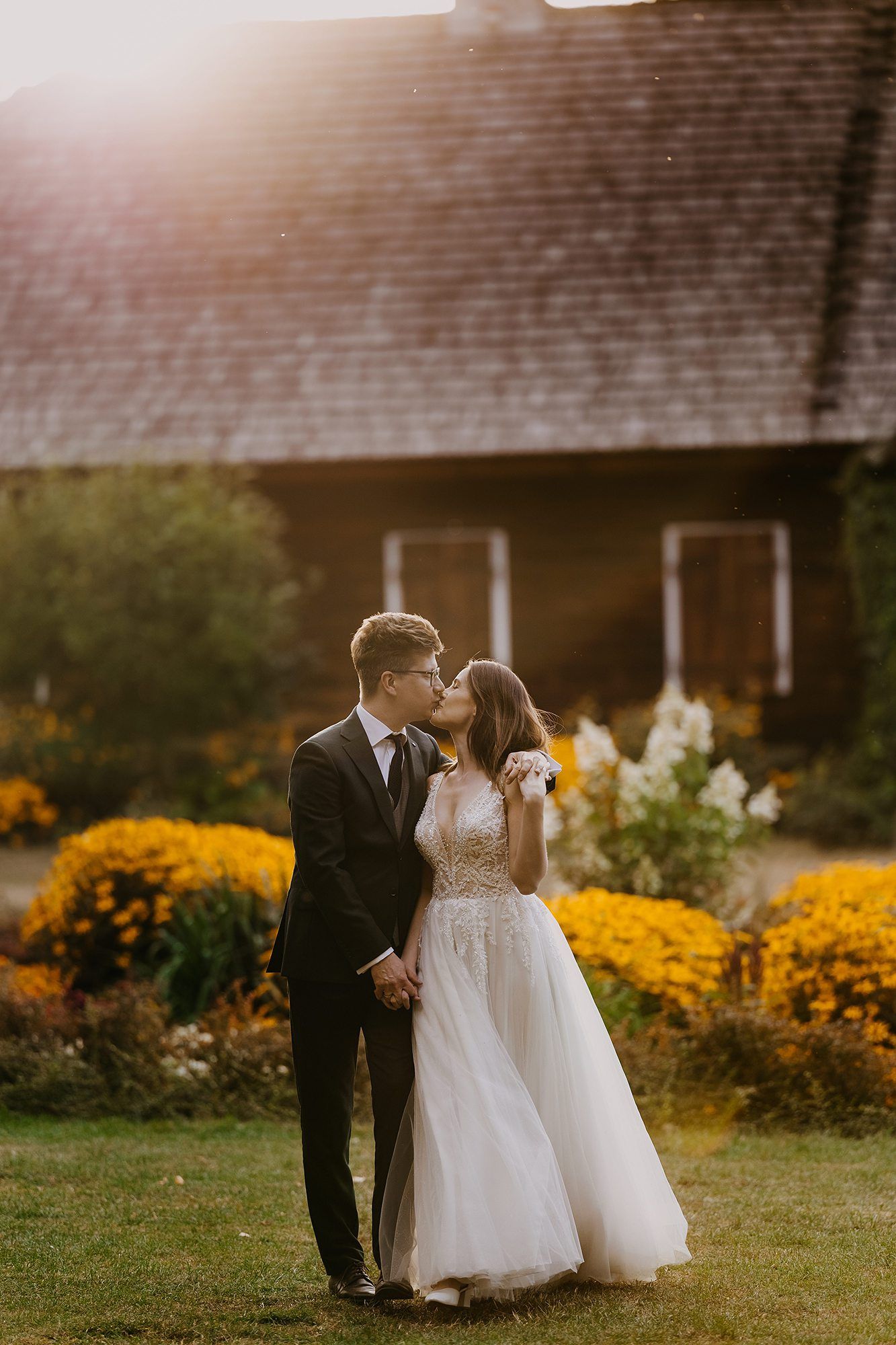 Bride and groom sharing a kiss at sunset in a garden with yellow flowers and rustic barn.