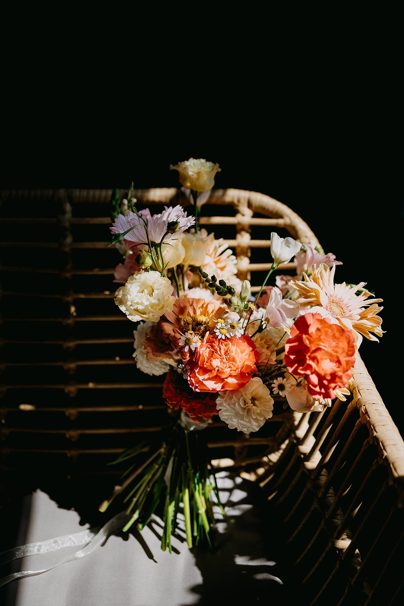 Vibrant bridal bouquet with orange, peach, and white flowers resting on a rattan chair in dramatic sunlight.