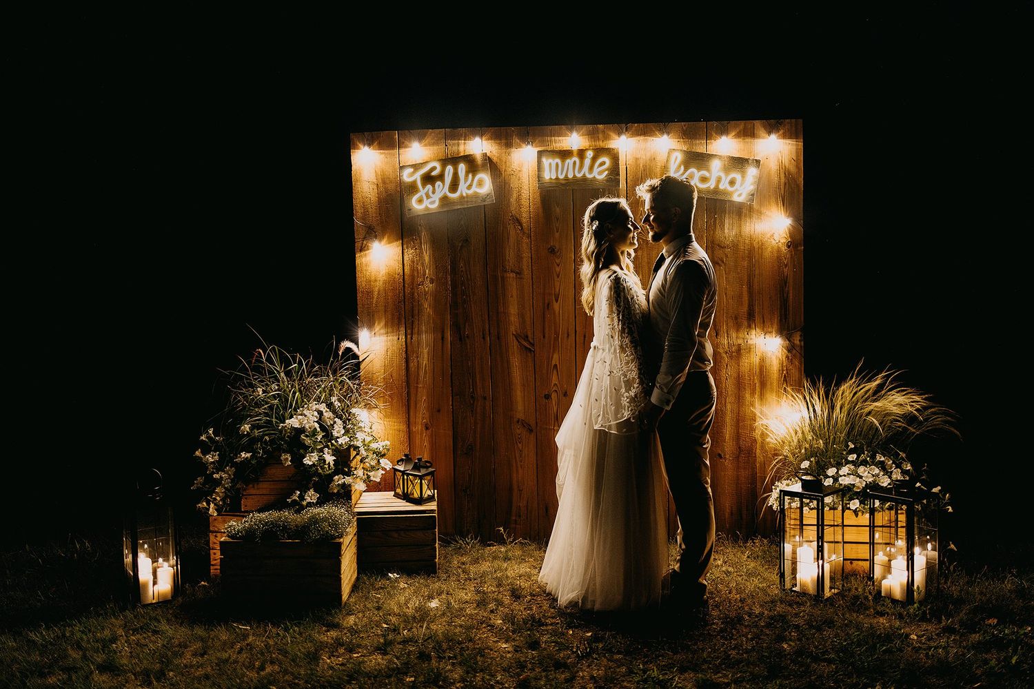 A romantic couple shares a kiss in front of a glowing wooden backdrop with fairy lights and floral arrangements at night.