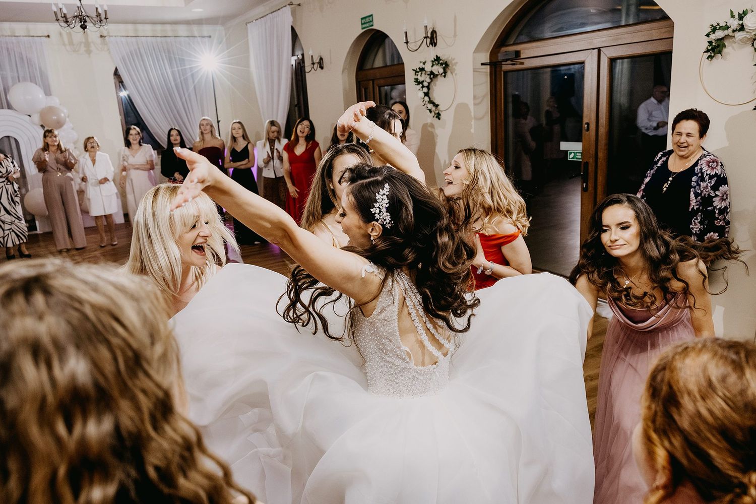 Bride dancing energetically with bridesmaids and guests in lace gown at wedding reception.