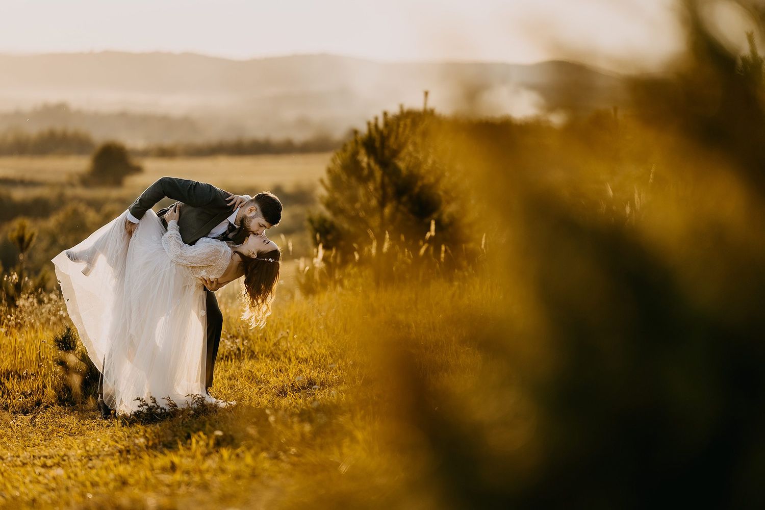 Groom dipping bride in romantic golden sunset embrace in scenic countryside meadow landscape.