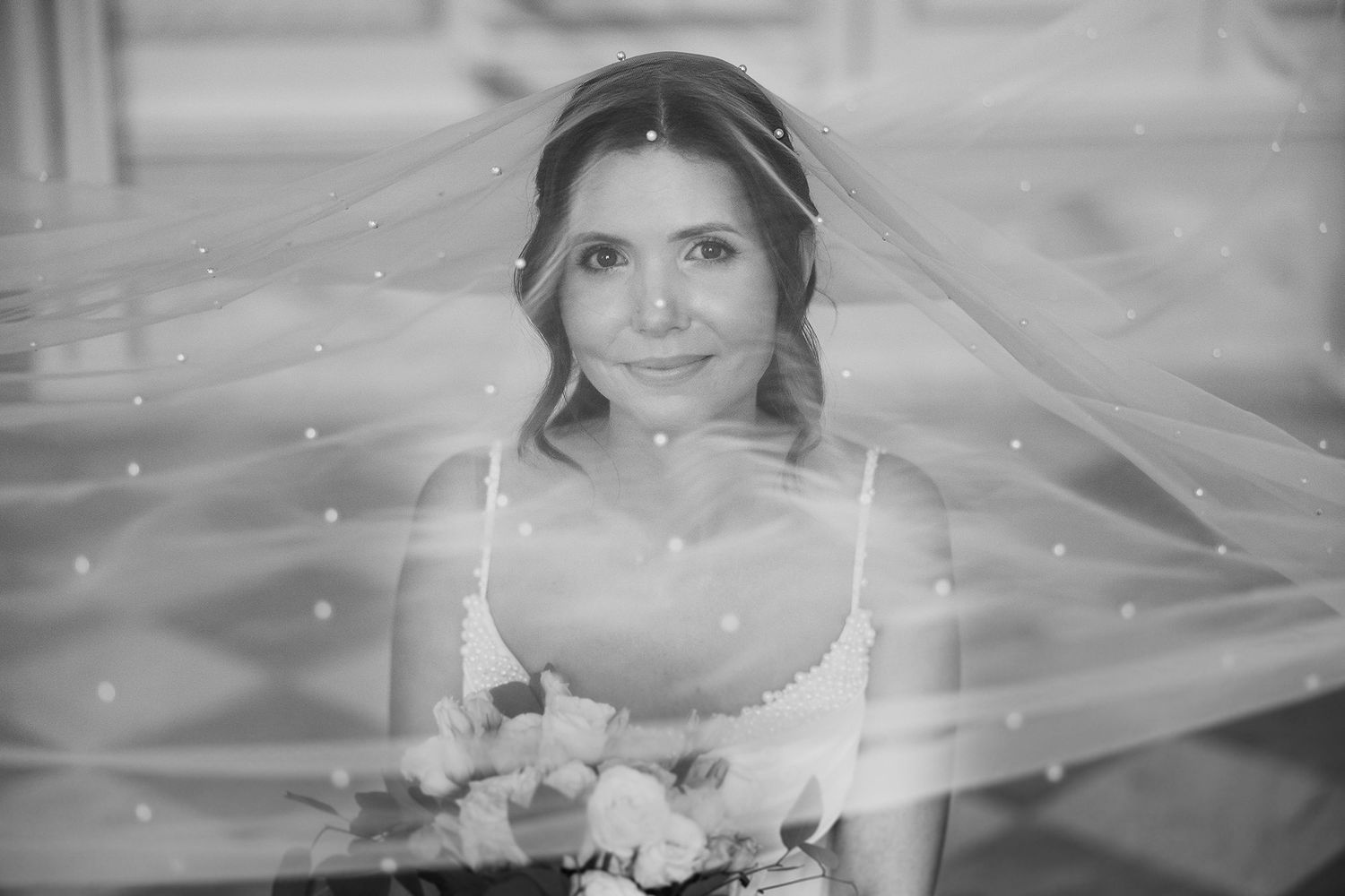 Black and white portrait of smiling bride beneath sheer veil with floral bouquet.