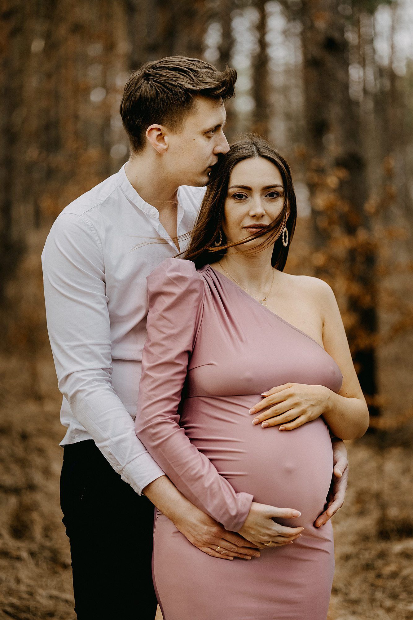 Man tenderly embracing pregnant woman in pink dress during outdoor autumn forest maternity shoot.