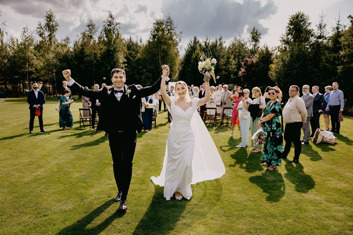 A joyful newlywed couple raises their hands in celebration while walking through guests on a green lawn.