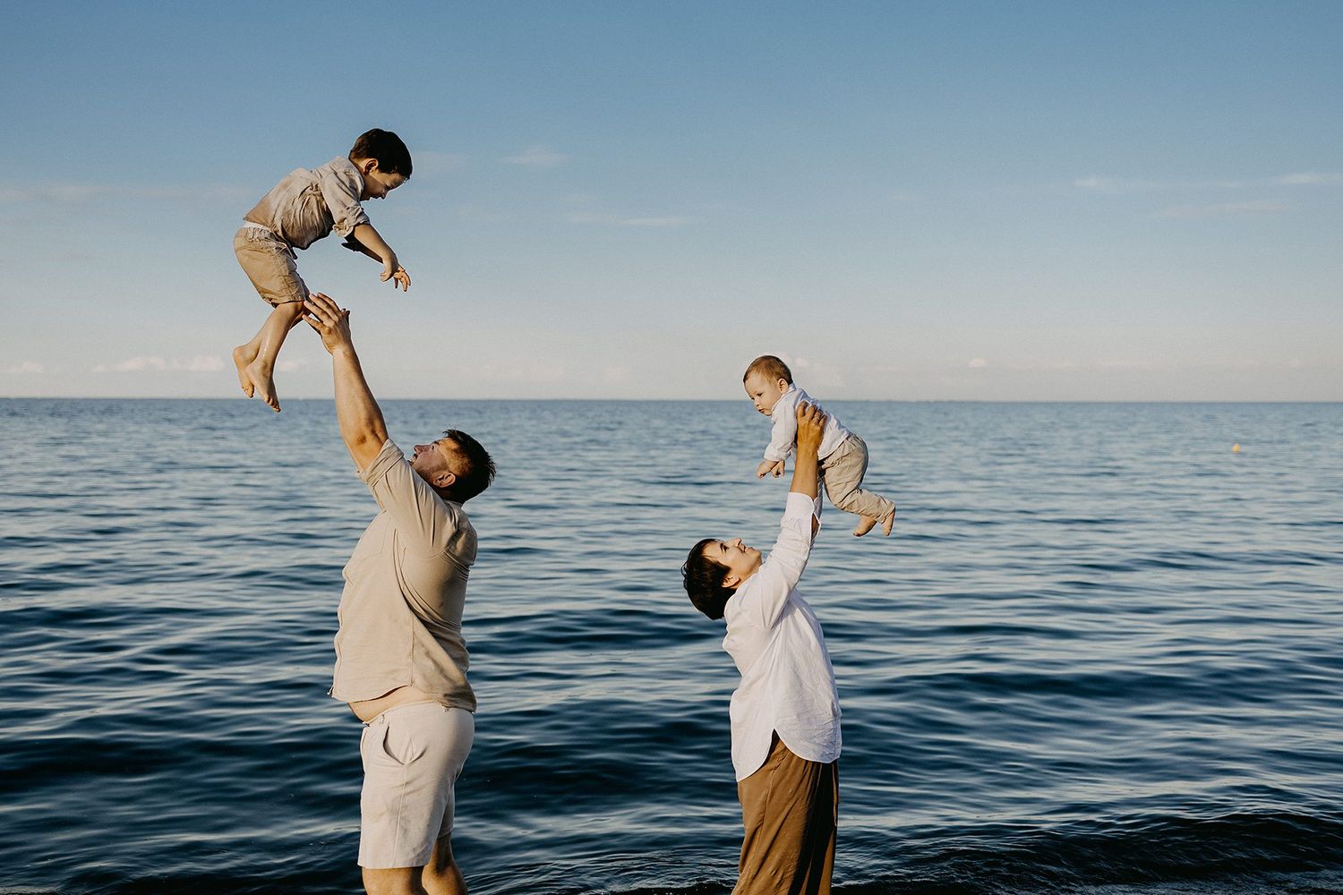 Parents lifting two young children joyfully above their heads while standing in calm blue ocean water.