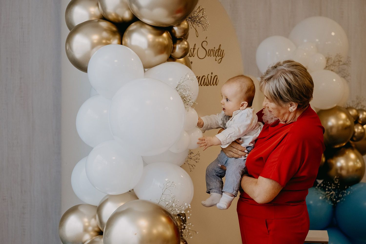 Grandmother holding baby near gold and white balloon arch at christening celebration party.