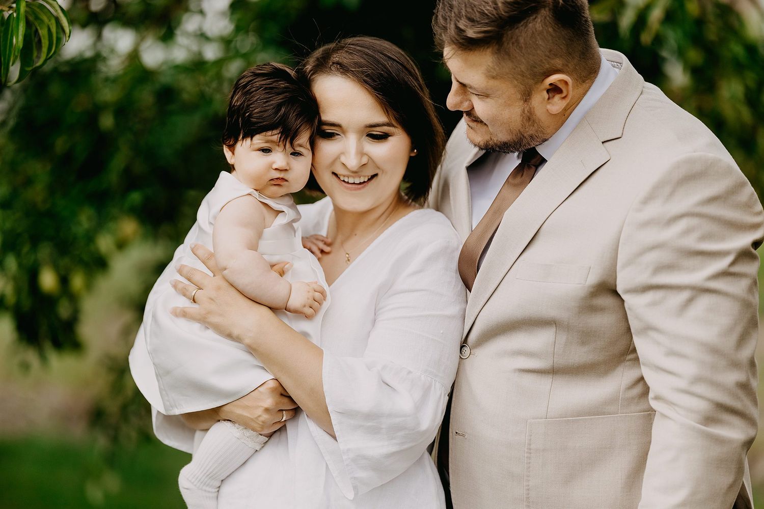 Happy young family with baby outdoors, mother in white dress and father in beige suit among greenery.