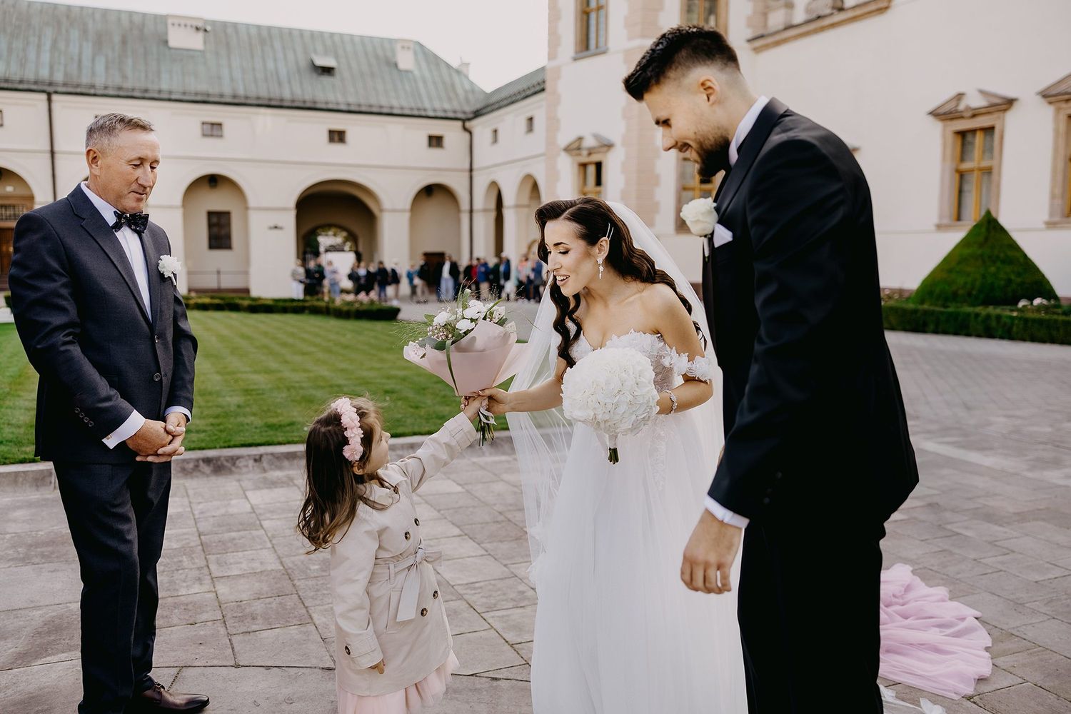 Bride and groom greeted by a young girl with flowers outside a historic European castle wedding venue.