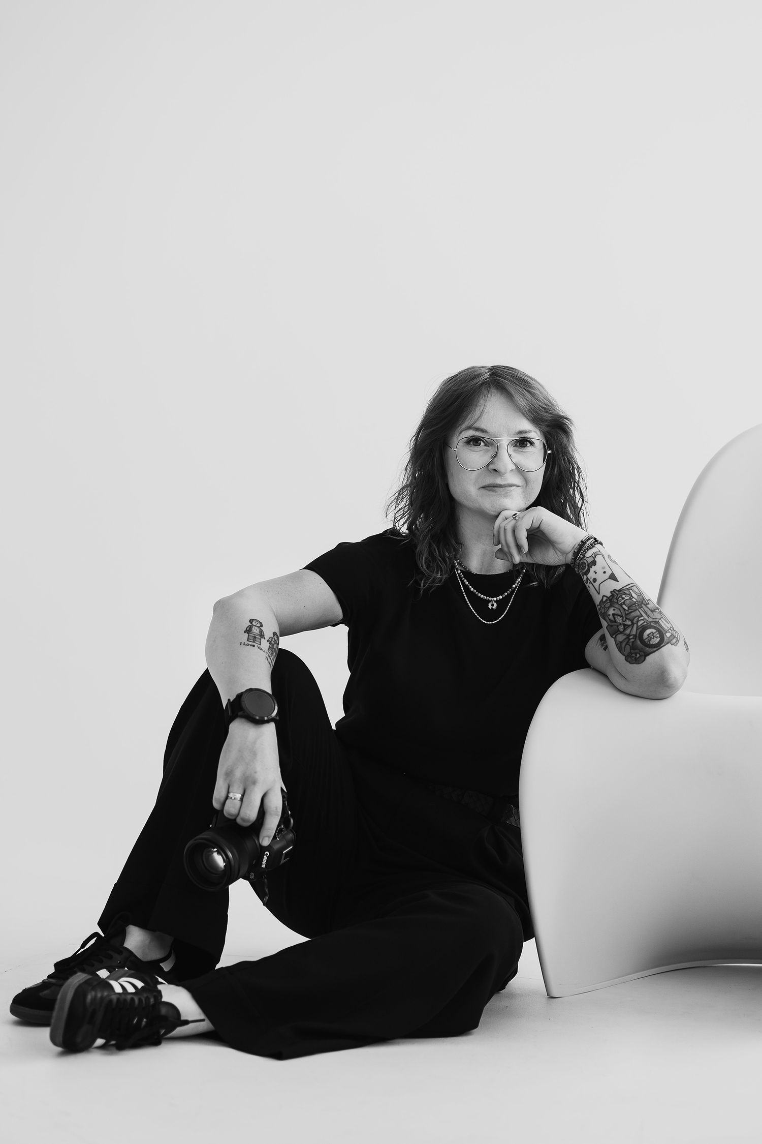 Black and white portrait of a tattooed woman in black outfit sitting relaxed against a modern white chair.