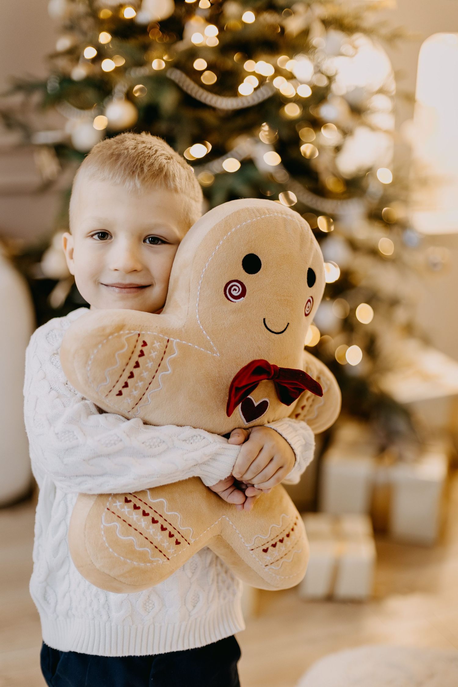 Smiling little boy in a white knit sweater hugs a plush gingerbread man pillow with a red bow near a glowing Christmas tree.