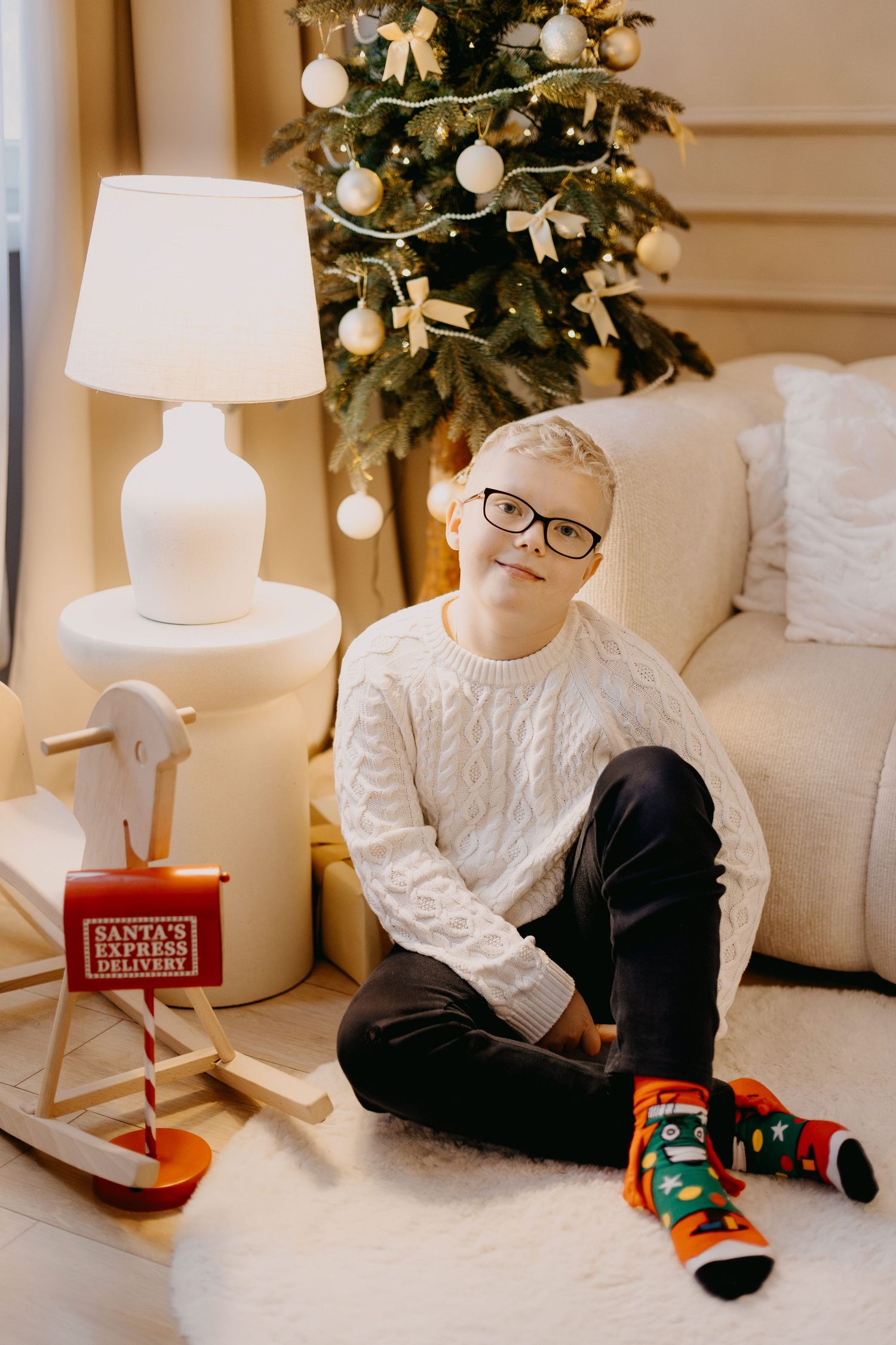 Smiling boy with glasses in a cream cable-knit sweater and festive Christmas socks sits beside a decorated tree and a Santa's Express toy.
