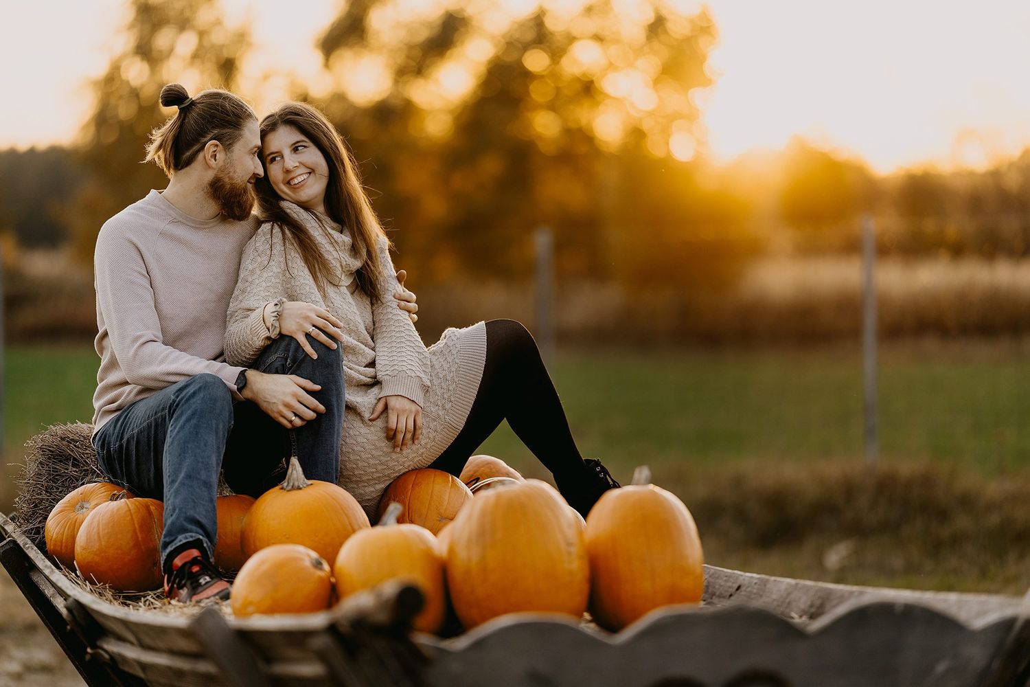 Couple sitting together on a wheelbarrow surrounded by orange pumpkins at golden hour.