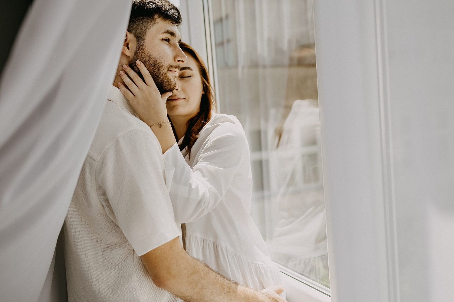 Romantic couple embracing tenderly by a sunlit window with sheer white curtains.
