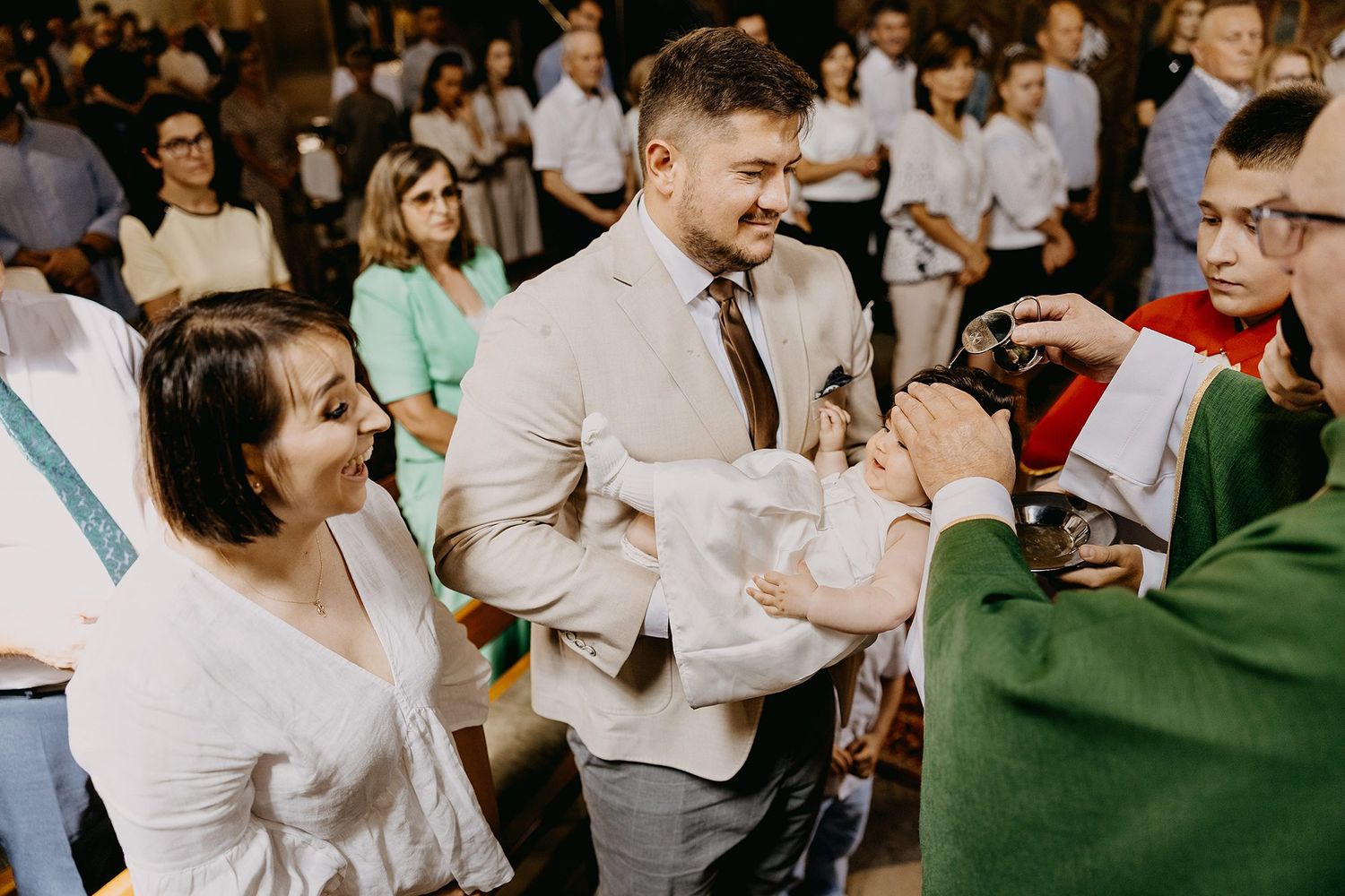 Smiling couple receiving baptism sacrament for their baby from priest during Catholic church ceremony.